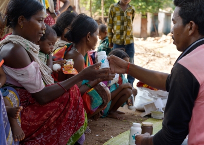 A line of women and children crouched on the ground in a line in India, while a man hands one woman a bottle of UNIMMAP MMS.
